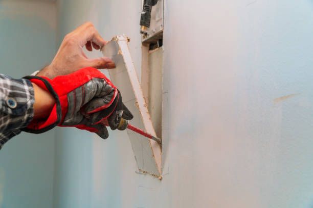 Worker cutting a rectangular drywall opening beside an electrical box