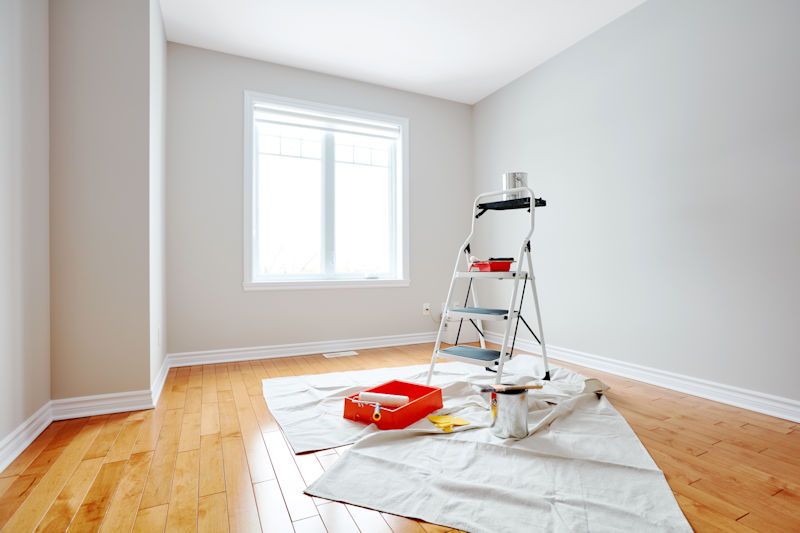Bright interior room with gray walls, a ladder, paint tray, and protected wood floor